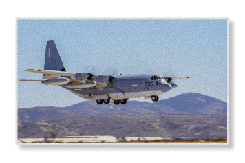 A gray C-130 Hercules military transport aircraft flies low over a desert airstrip with mountains in the background.