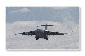 A C-17 Globemaster III military transport aircraft on final approach with landing gear deployed against a cloudy sky.