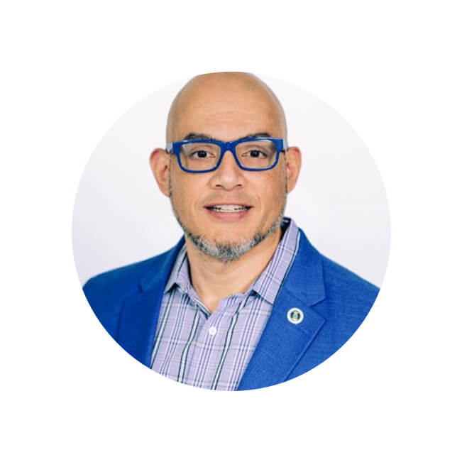 Omar Velasco, a bald man with a goatee wearing blue glasses, a plaid shirt, and a blue blazer, smiling warmly.