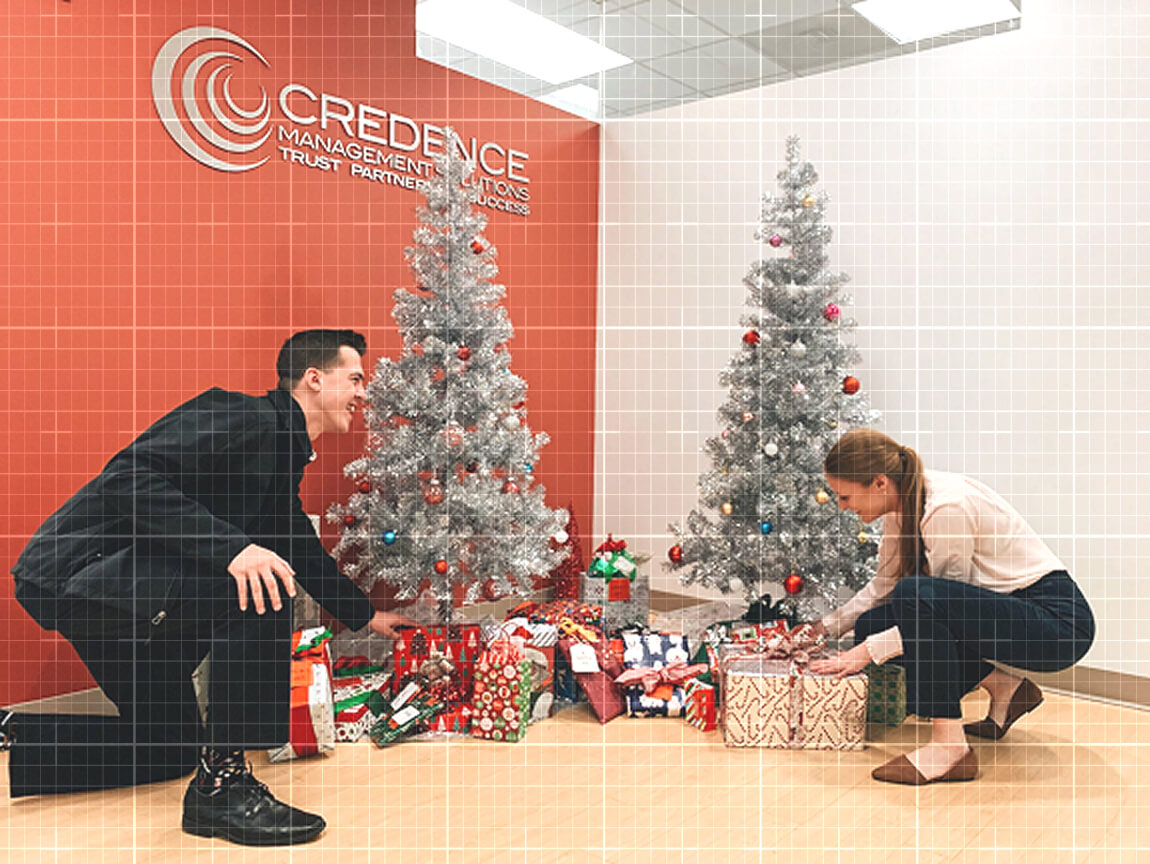 Two Credence Management Solutions employees arrange wrapped gifts beneath silver Christmas trees in the office lobby.