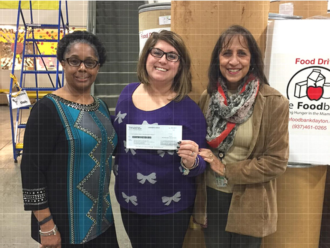 Three smiling women holding a donation check at a food bank during a community food drive event.