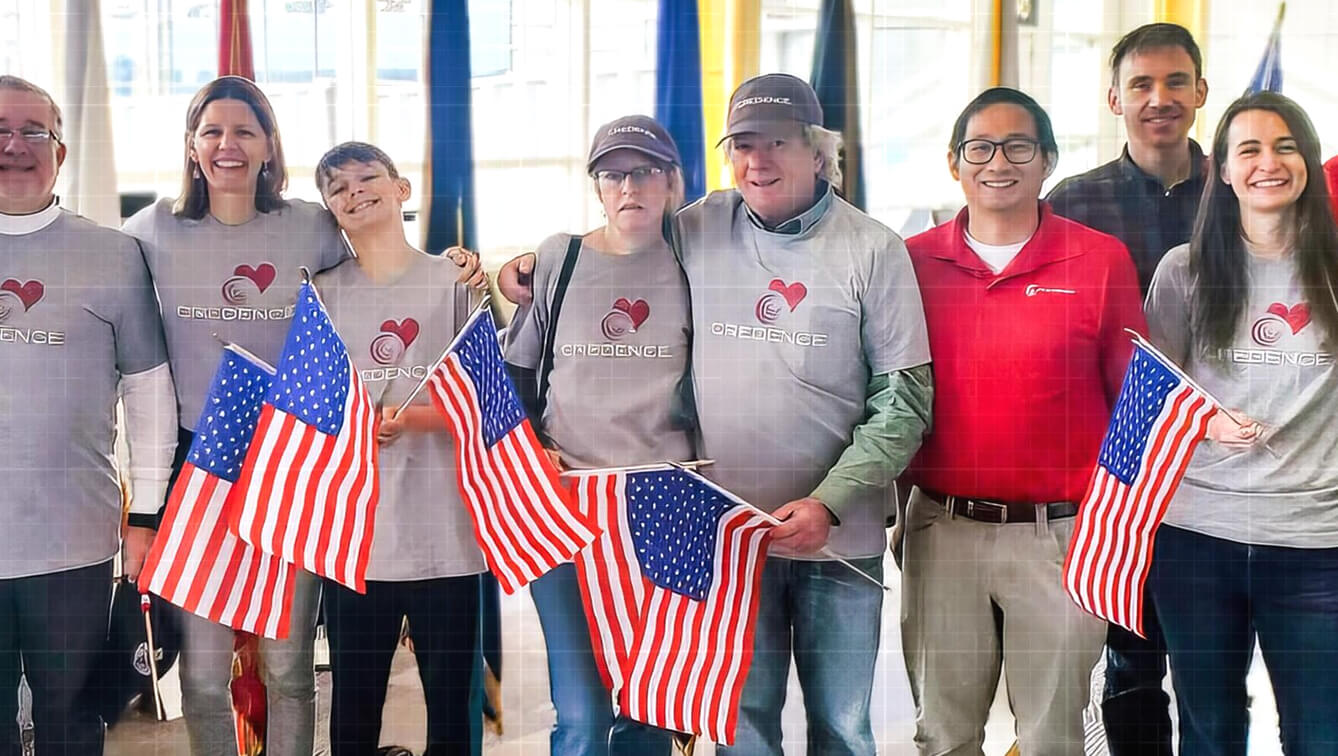 Smiling Credence team members wearing branded shirts pose together holding small American flags at a company event.