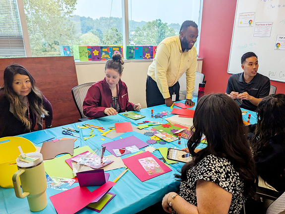 Volunteers gather around a table covered with colorful paper and craft supplies, creating handmade cards for hospitalized