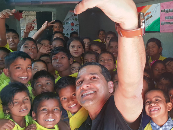 A smiling man takes a selfie surrounded by a large group of happy children in yellow uniforms at a community event.