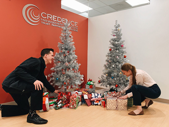 Two employees arrange wrapped gifts beneath silver Christmas trees in the Credence Management Solutions office lobby.