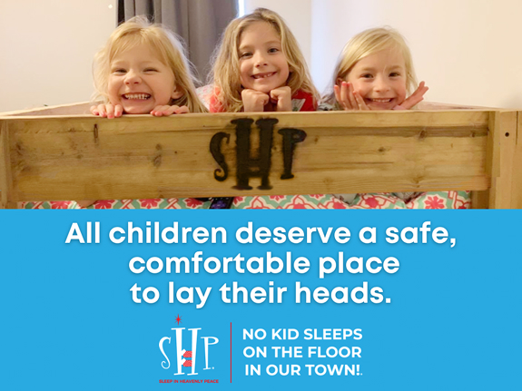Three smiling young girls peek over a wooden bed branded with the Sleep in Heavenly Peace logo and mission message.