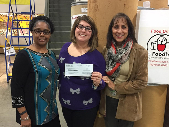 Three smiling women pose together holding a donation check at The Dayton Foodbank warehouse during a food drive event.
