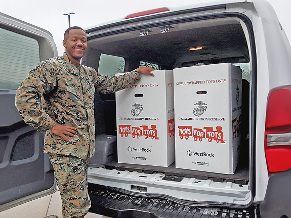 A smiling U.S. Marine in camouflage uniform stands beside a vehicle loaded with Toys for Tots donation boxes.