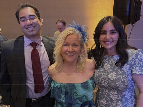 Three smiling team members posing together at a formal event, with the women in floral dresses and the man in a suit and tie.