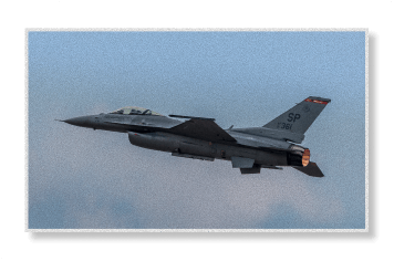 An F-16 fighter jet marked "SP" flies through an overcast sky with its afterburner glowing orange.