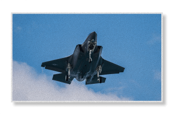 An F-35 stealth fighter jet flies overhead against a blue sky with white clouds below it.