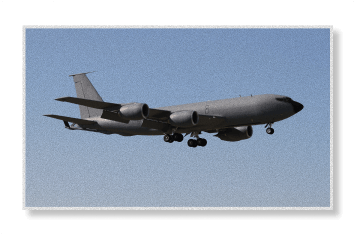 A gray KC-135 Stratotanker military aerial refueling aircraft in flight with landing gear deployed against a clear sky.