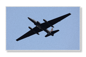 A U-2 reconnaissance aircraft flies overhead against a clear blue sky, its long slender wings silhouetted in dark profile.